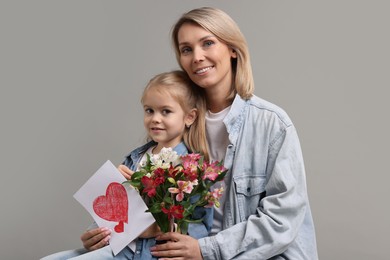 Photo of Happy woman with her daughter, bouquet of alstroemeria flowers and greeting card on gray background. Mother's Day celebration