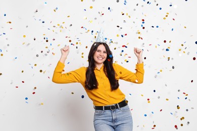 Photo of Happy woman in conical paper hat under falling confetti on white background. Surprise party