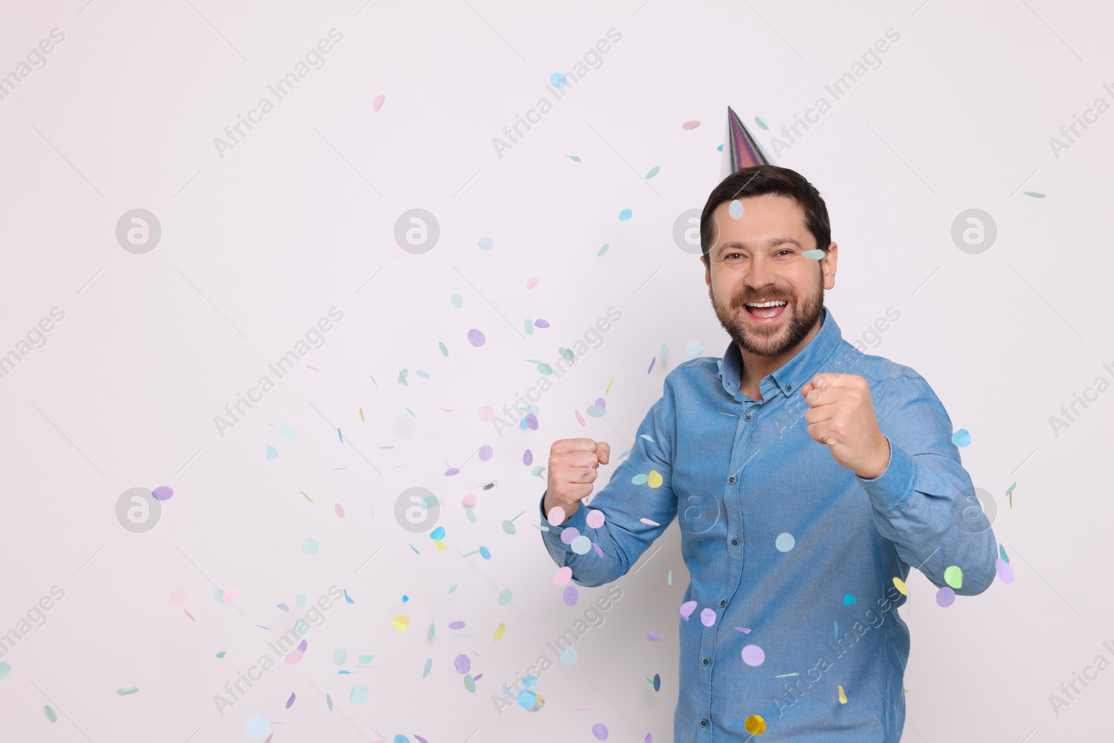 Photo of Happy man in conical paper hat and falling confetti on white background, space for text. Surprise party