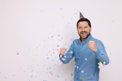 Photo of Happy man in conical paper hat and falling confetti on white background, space for text. Surprise party