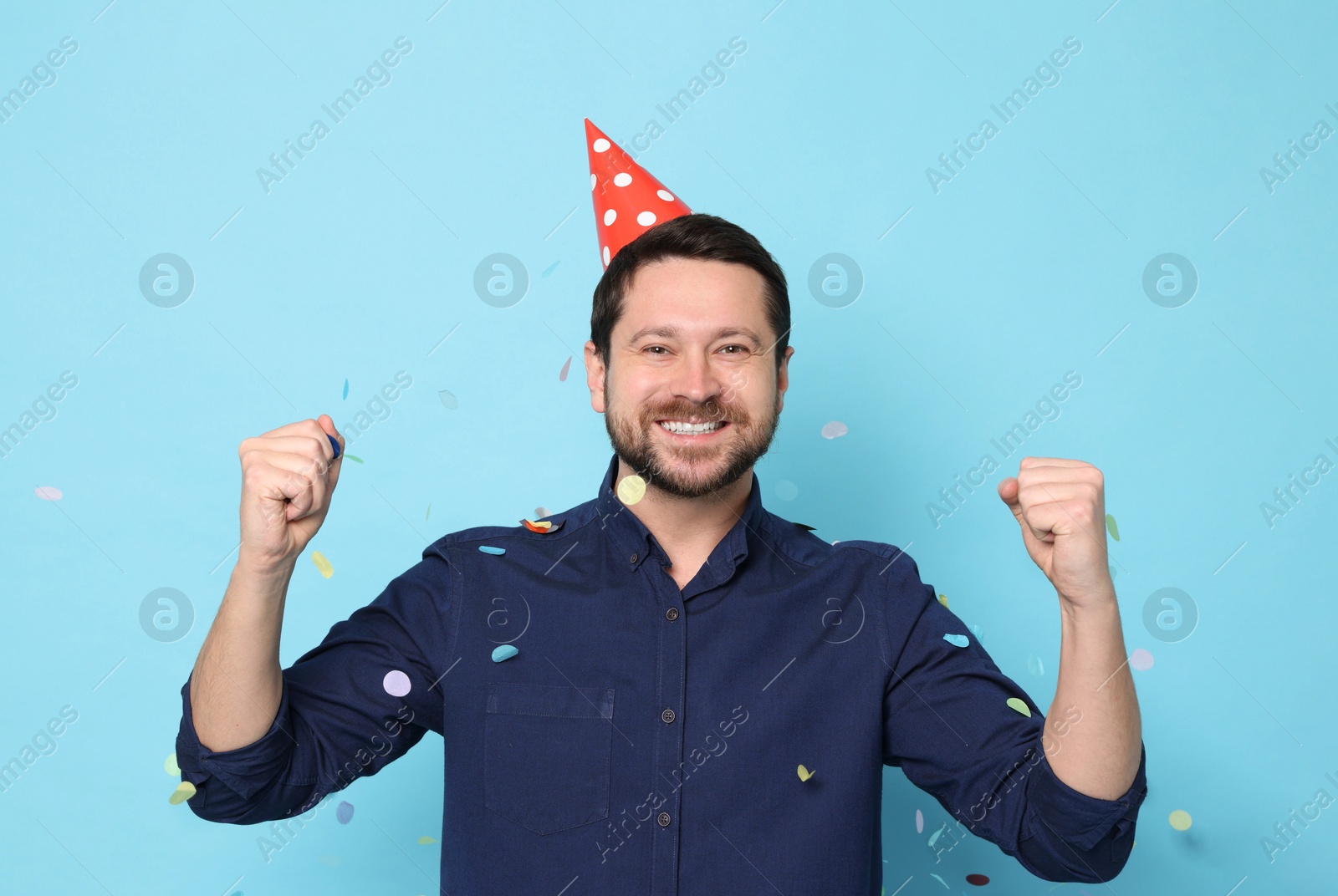 Photo of Happy man in conical paper hat and flying confetti on light blue background. Surprise party