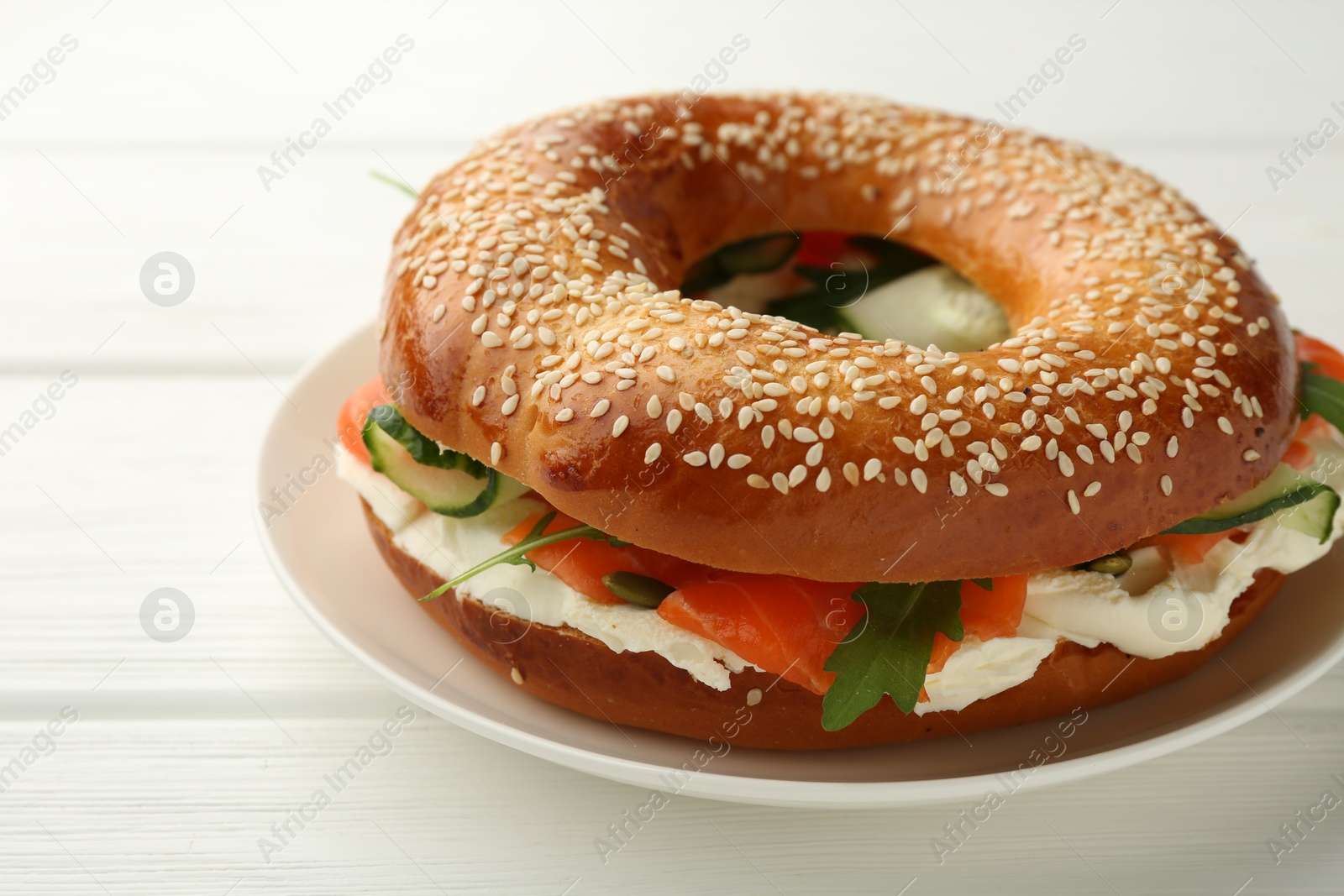 Photo of Delicious bagel with salmon, cream cheese and cucumber on white wooden table, closeup