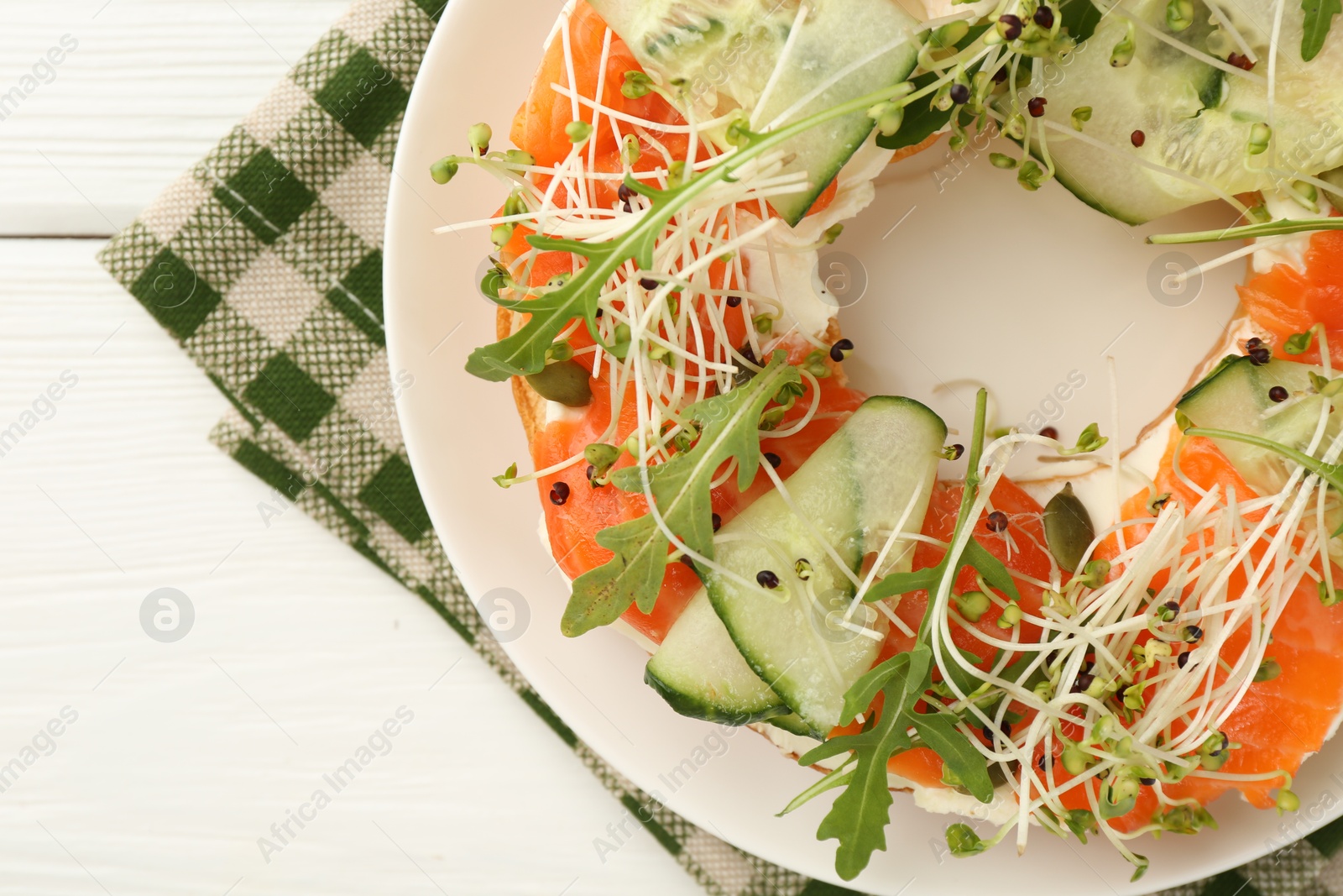 Photo of Half of delicious bagel with salmon, microgreens, arugula and cucumber on white wooden table, top view