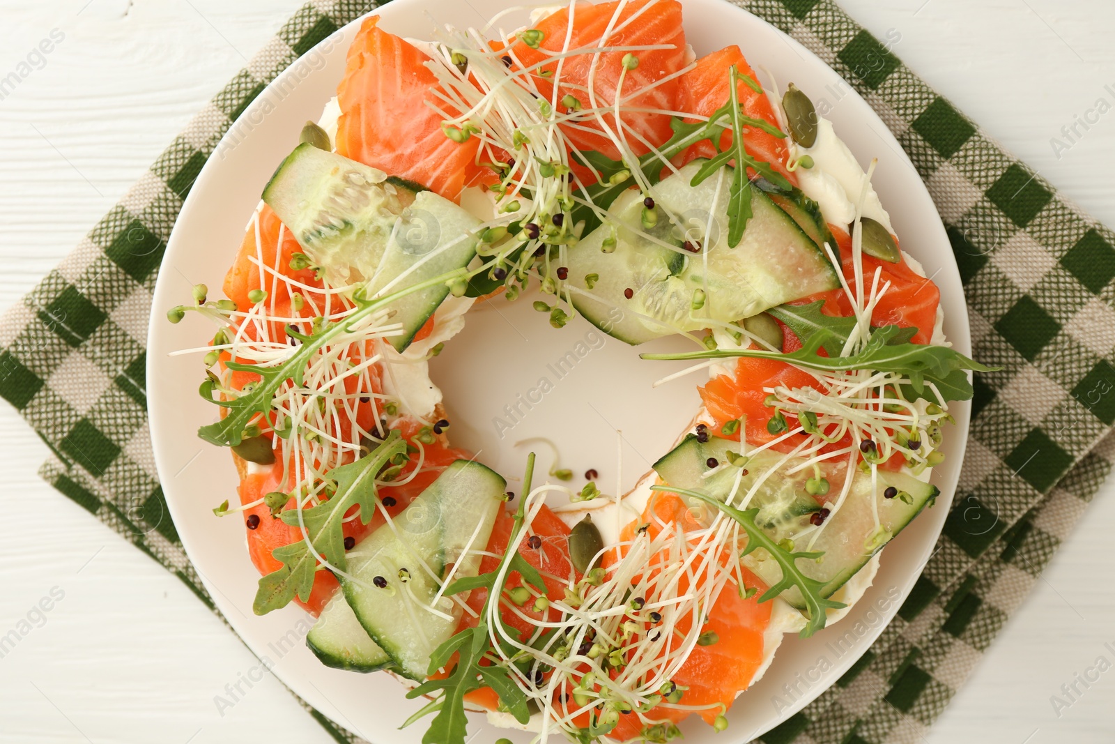 Photo of Half of delicious bagel with salmon, microgreens, arugula and cucumber on white wooden table, top view