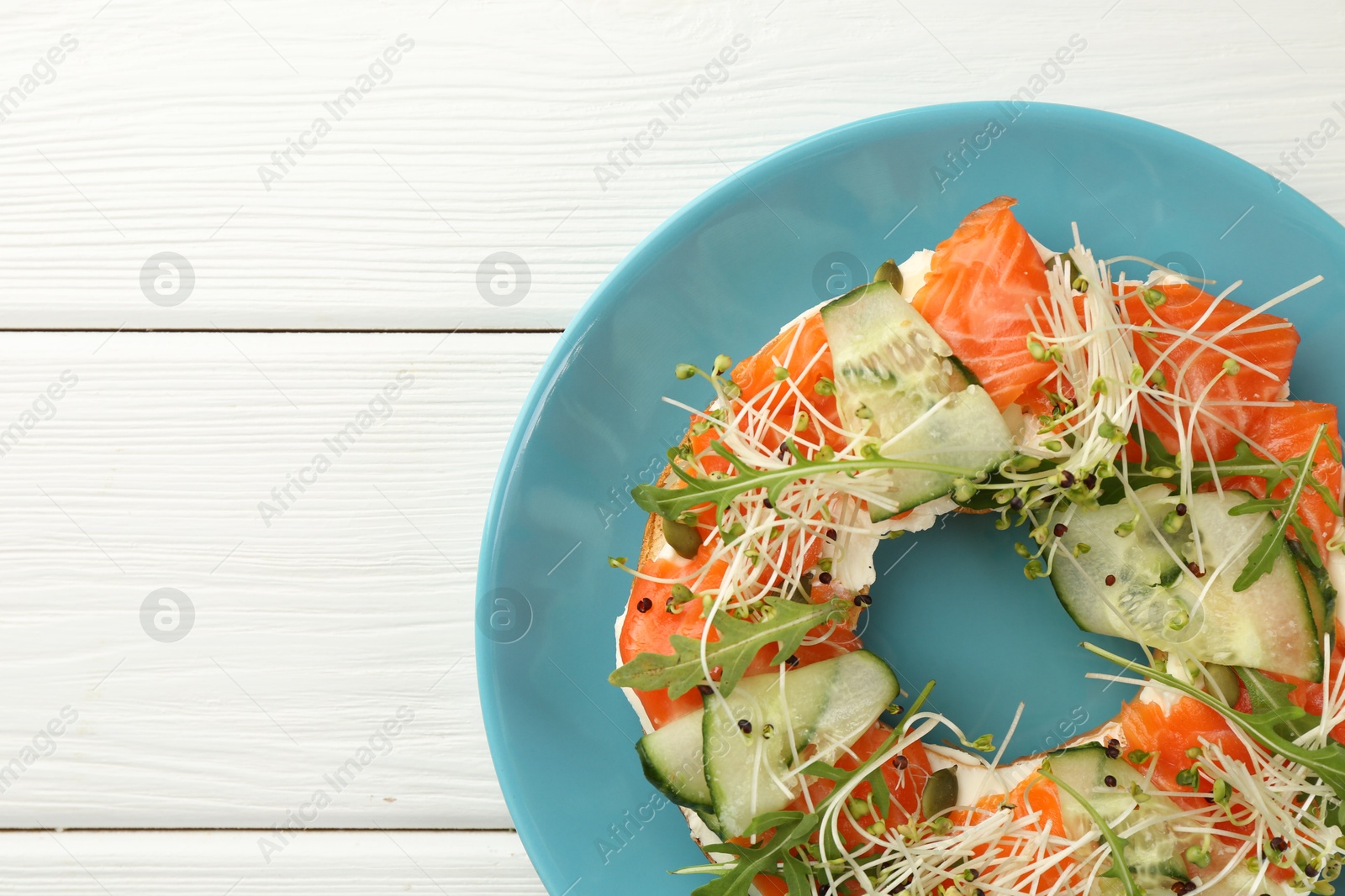 Photo of Half of delicious bagel with salmon, microgreens, arugula and cucumber on white wooden table, top view. Space for text