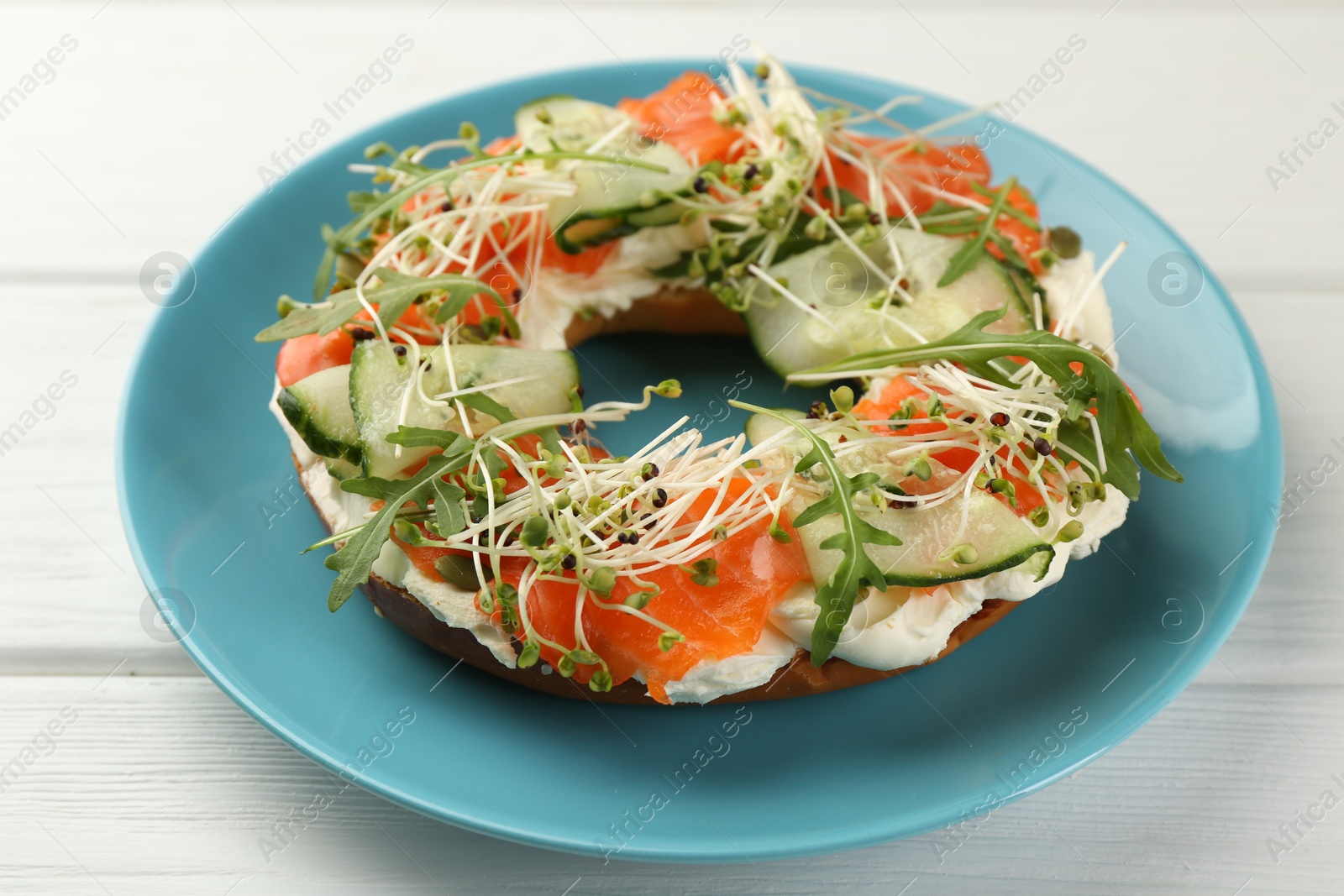 Photo of Half of delicious bagel with salmon, microgreens, arugula and cream cheese on white wooden table, closeup