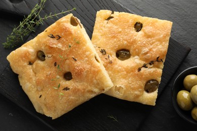 Photo of Pieces of delicious focaccia bread with olives and thyme on black table, flat lay