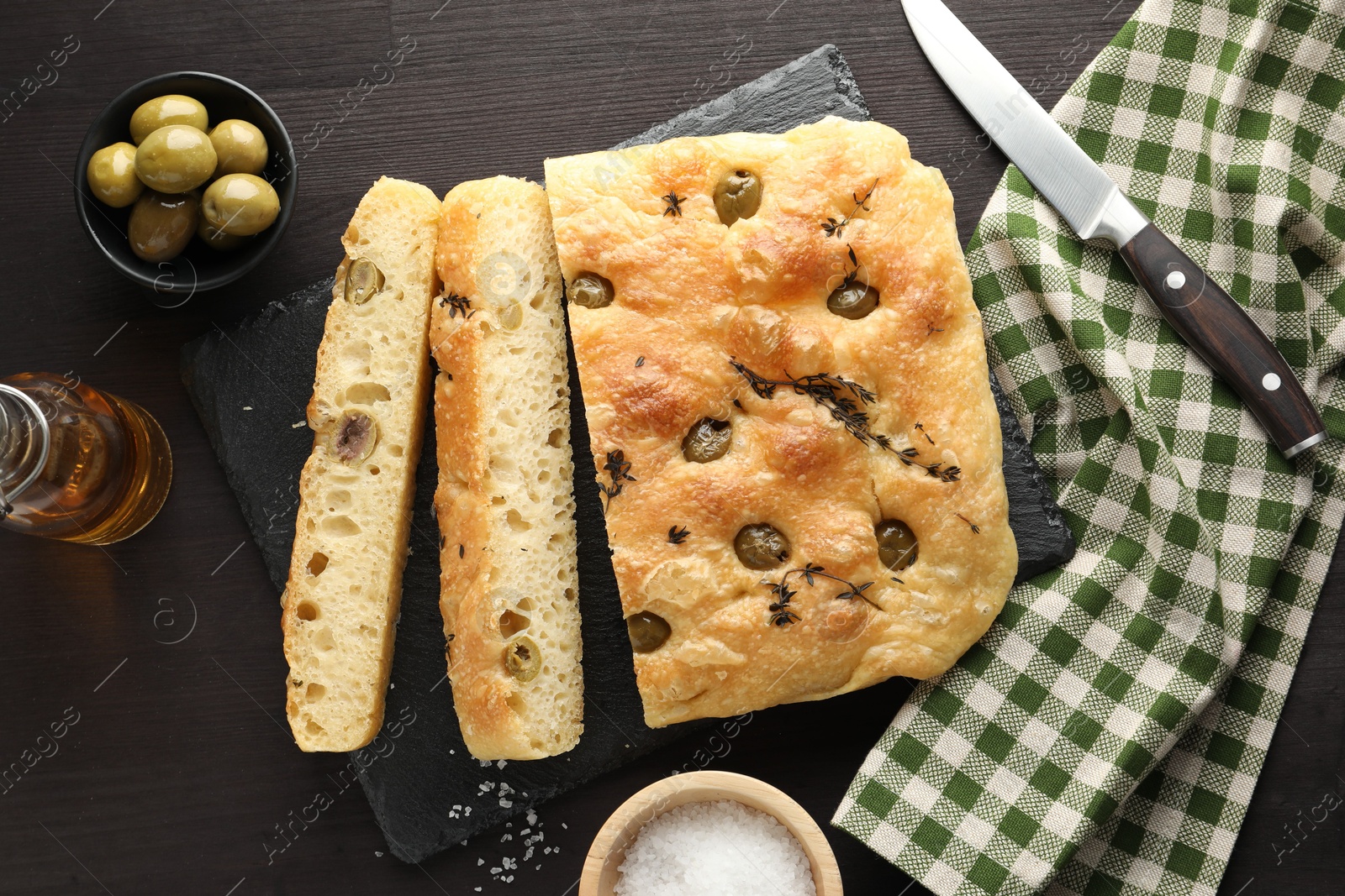 Photo of Pieces of delicious focaccia bread with olives, thyme, salt and knife on dark wooden table, flat lay