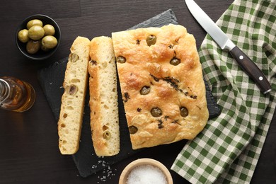 Photo of Pieces of delicious focaccia bread with olives, thyme, salt and knife on dark wooden table, flat lay