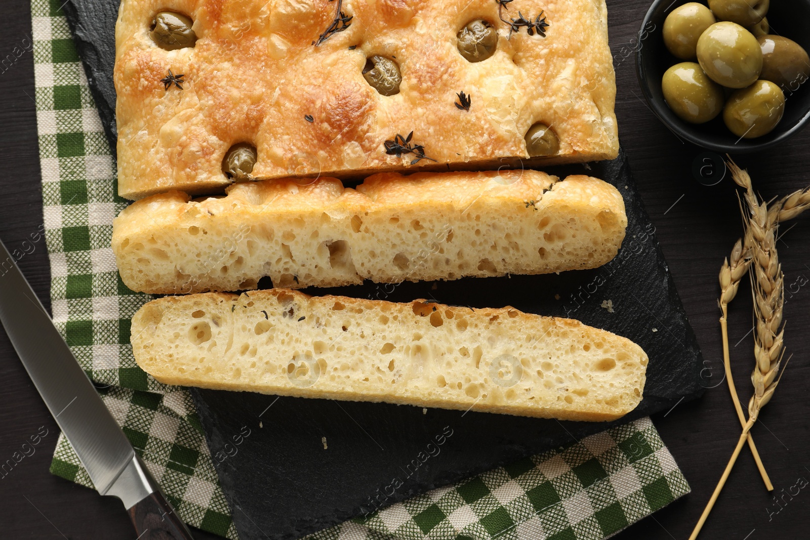 Photo of Pieces of delicious focaccia bread with olives, thyme, spikes and knife on dark wooden table, flat lay