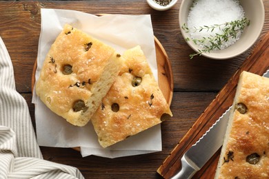 Photo of Pieces of delicious focaccia bread with olives, thyme, salt and knife on wooden table, flat lay