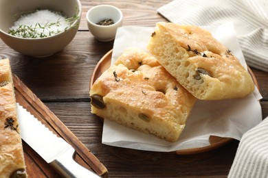 Photo of Pieces of delicious focaccia bread with olives, thyme, salt and knife on wooden table, closeup