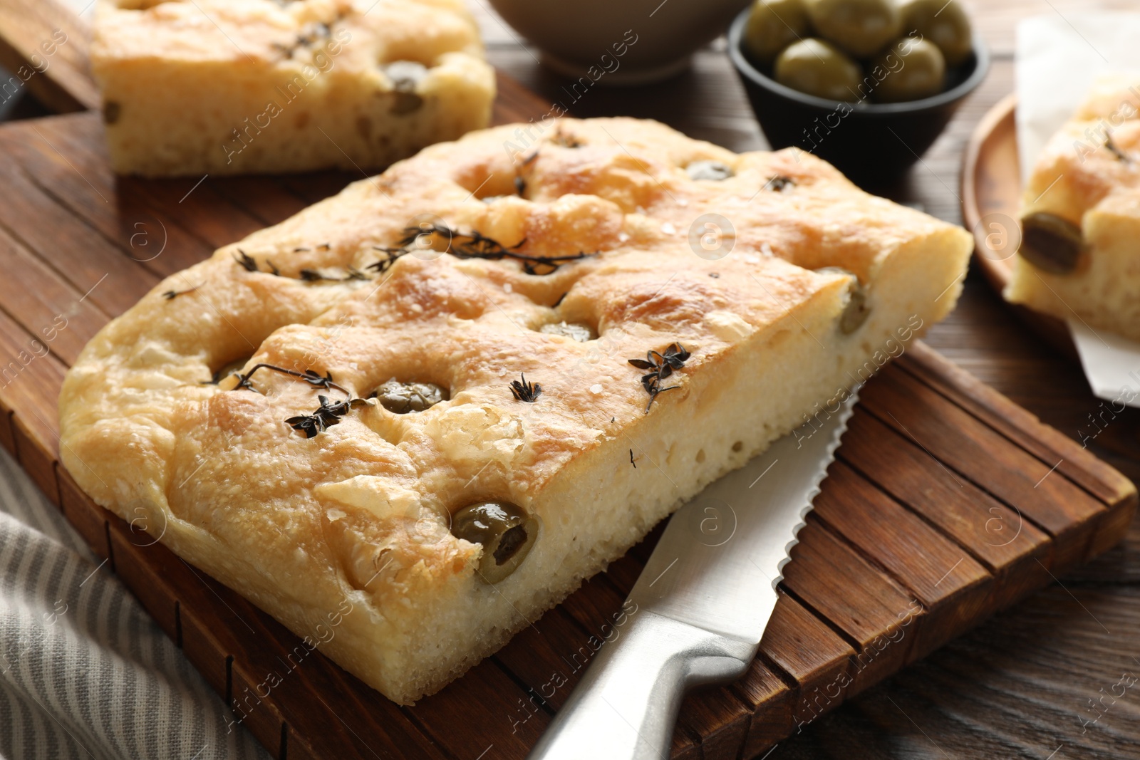 Photo of Pieces of delicious focaccia bread with olives, thyme, salt and knife on wooden table, closeup