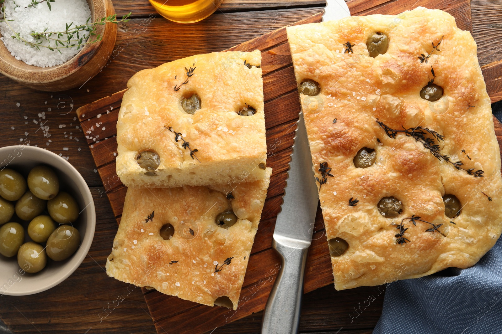 Photo of Pieces of delicious focaccia bread with olives, thyme, salt and knife on wooden table, flat lay