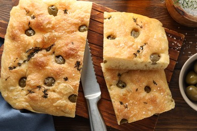 Photo of Pieces of delicious focaccia bread with olives, thyme, salt and knife on wooden table, flat lay