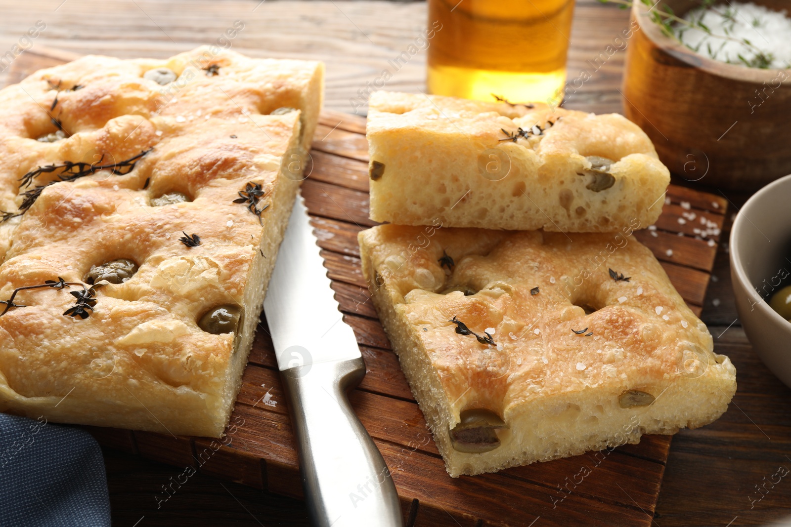 Photo of Pieces of delicious focaccia bread with olives, thyme, salt and knife on wooden table, closeup