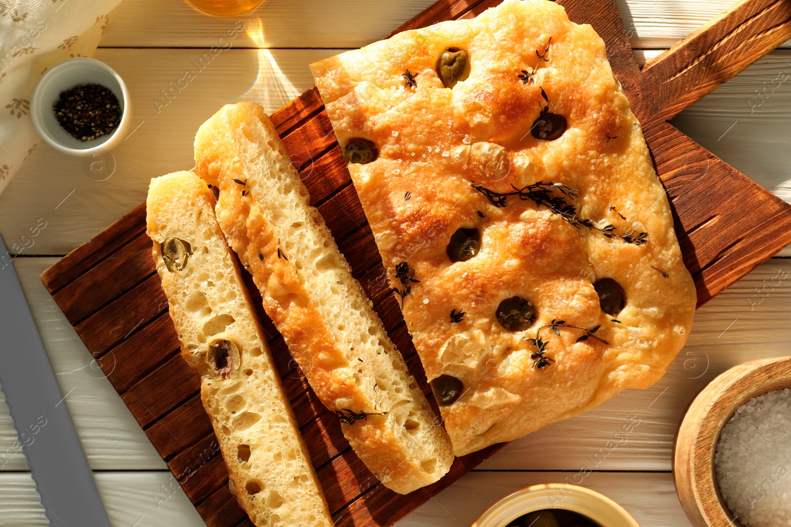 Photo of Pieces of delicious focaccia bread with olives, thyme, spices and knife on white wooden table, flat lay