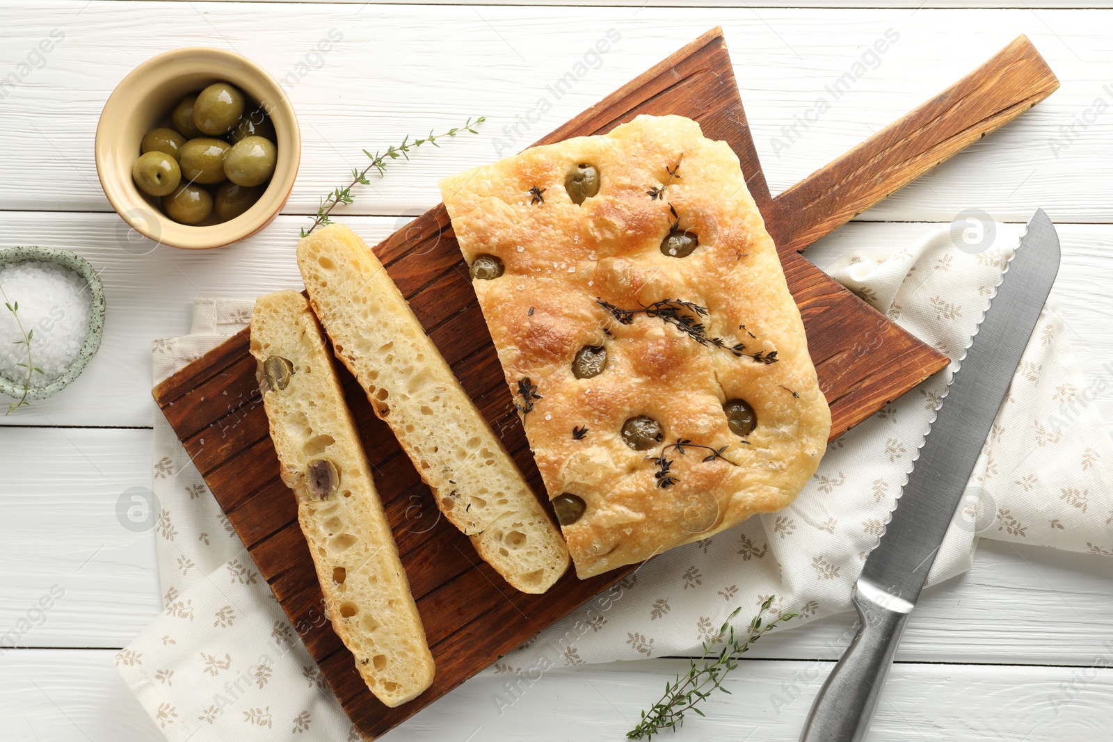 Photo of Pieces of delicious focaccia bread with olives, thyme, salt and knife on white wooden table, flat lay