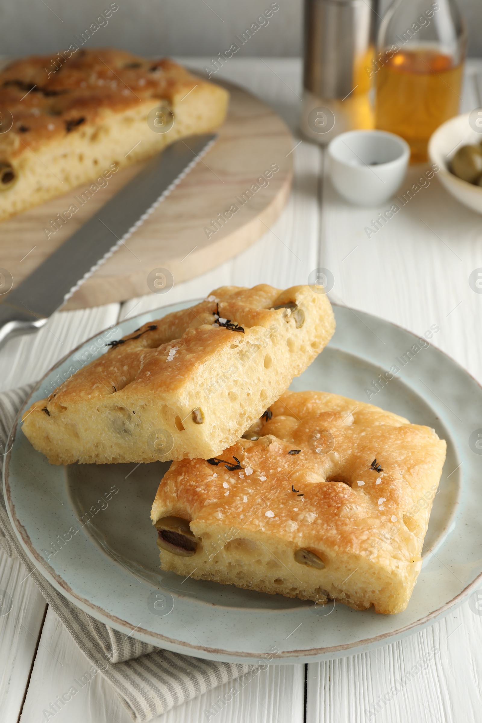 Photo of Pieces of delicious focaccia bread with olives, thyme, salt and knife on white wooden table, closeup