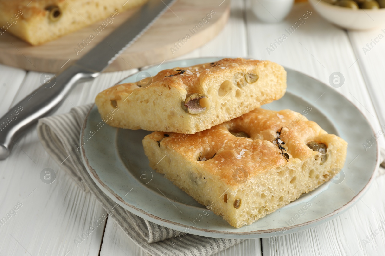 Photo of Pieces of delicious focaccia bread with olives, thyme, salt and knife on white wooden table, closeup