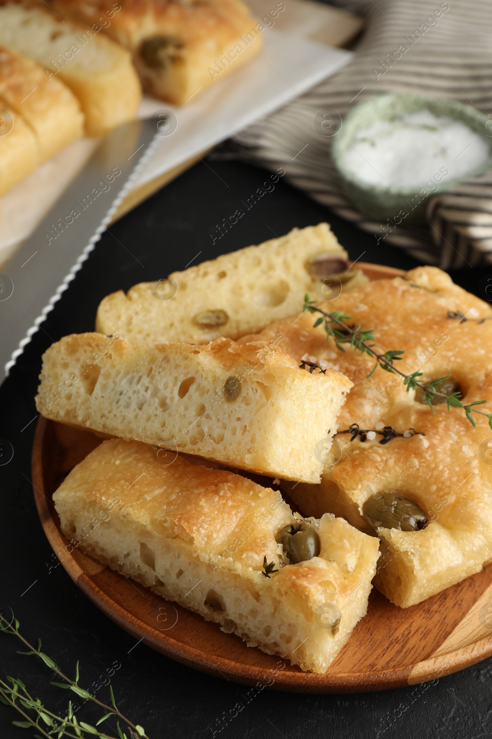 Photo of Slices of delicious focaccia bread with olives, thyme and salt on black table, closeup