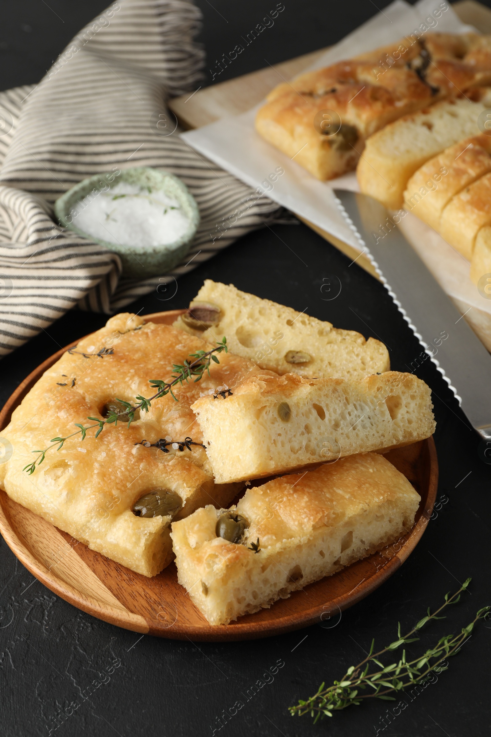 Photo of Slices of delicious focaccia bread with olives, thyme, salt and knife on black table, closeup