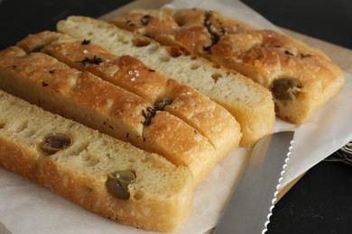 Photo of Slices of delicious focaccia bread with olives, thyme, salt and knife on black table, closeup