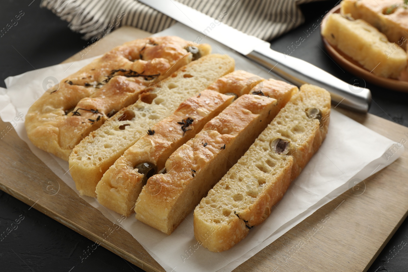 Photo of Slices of delicious focaccia bread with olives, thyme, salt and knife on black table, closeup