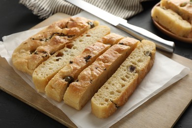 Photo of Slices of delicious focaccia bread with olives, thyme, salt and knife on black table, closeup