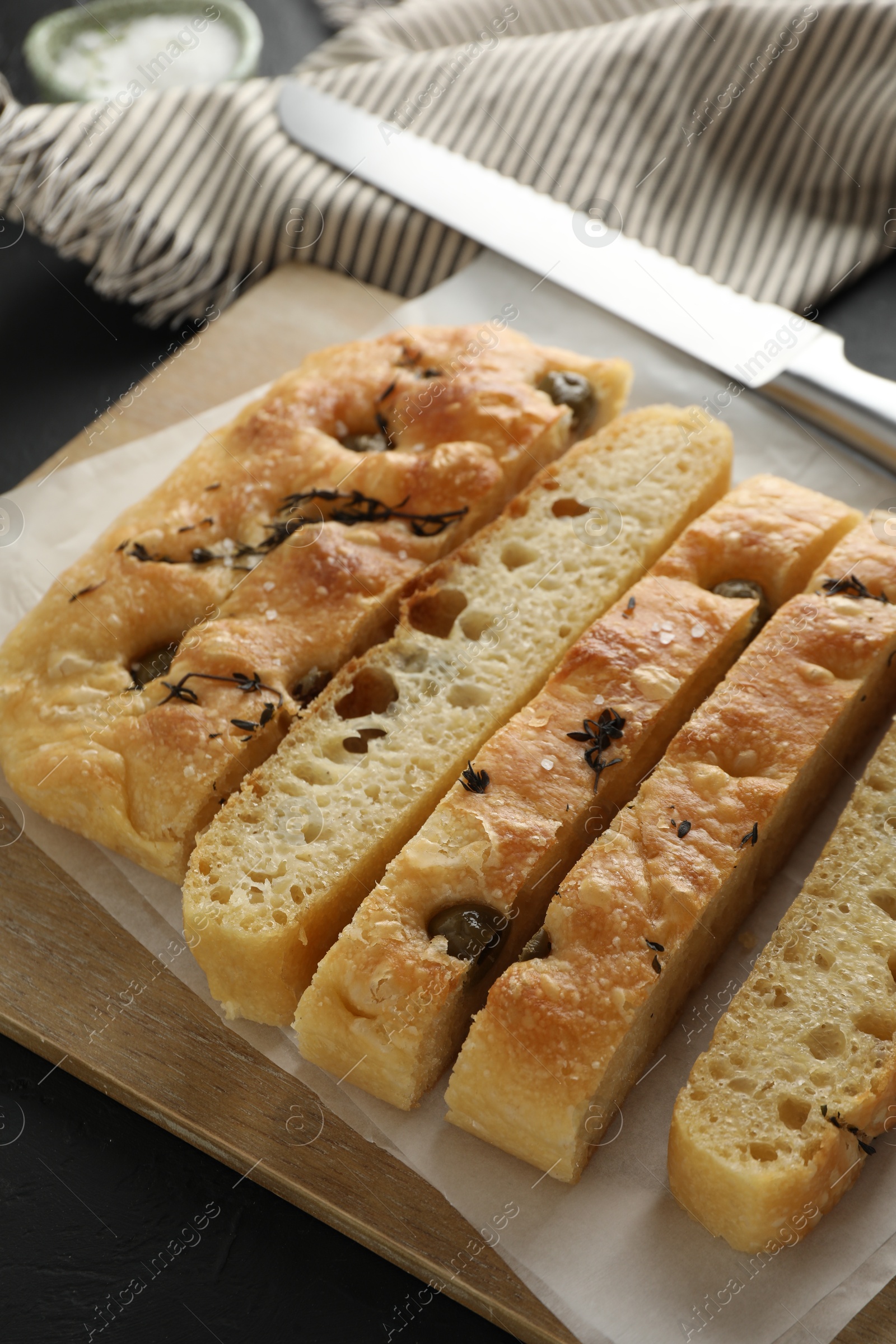 Photo of Slices of delicious focaccia bread with olives, thyme, salt and knife on black table, closeup