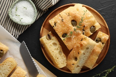 Photo of Slices of delicious focaccia bread with olives, thyme, salt and knife on black table, flat lay