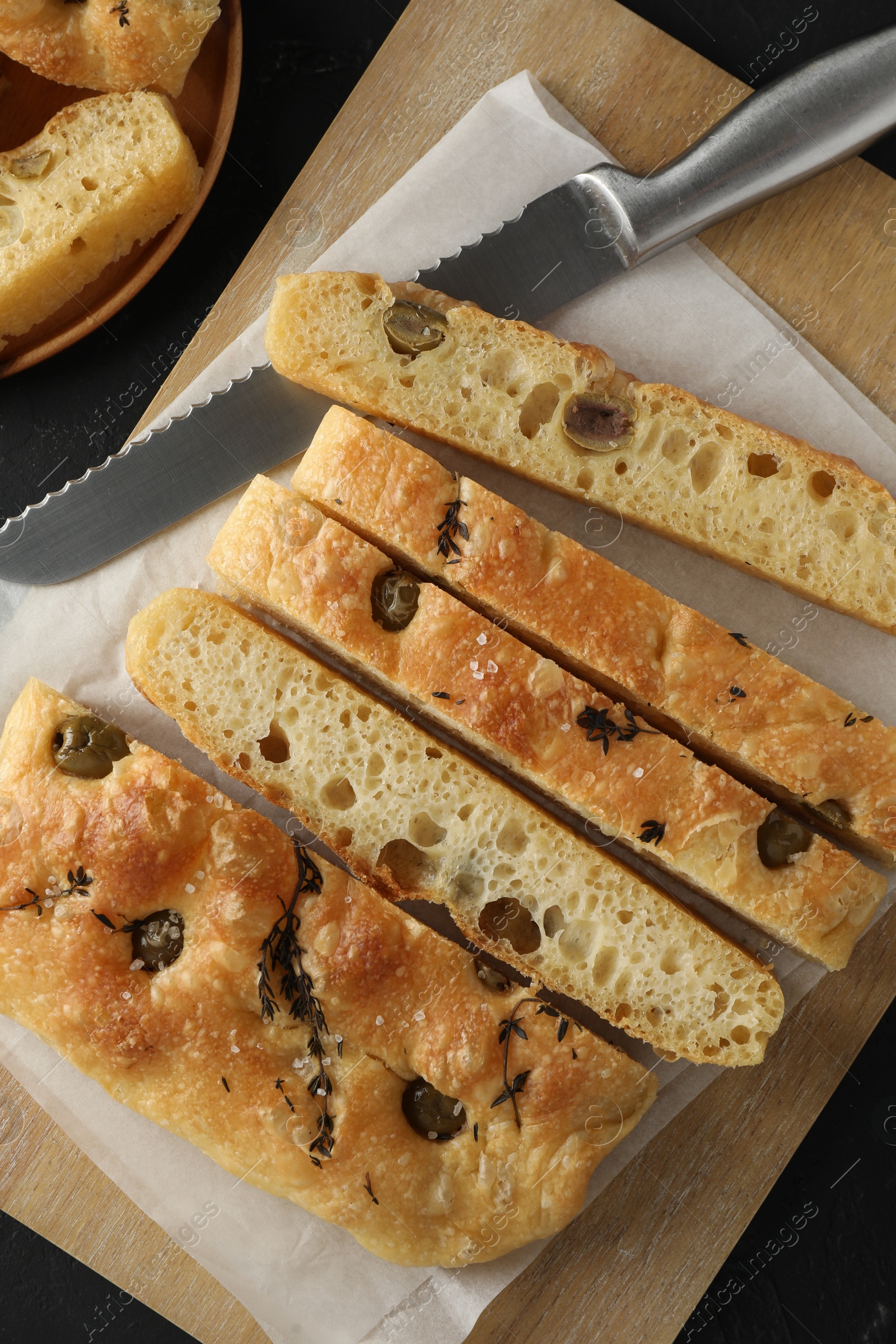 Photo of Slices of delicious focaccia bread with olives, thyme, salt and knife on black table, top view