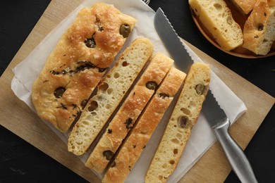 Photo of Slices of delicious focaccia bread with olives, thyme, salt and knife on black table, flat lay