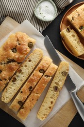 Photo of Slices of delicious focaccia bread with olives, thyme, salt and knife on black table, flat lay