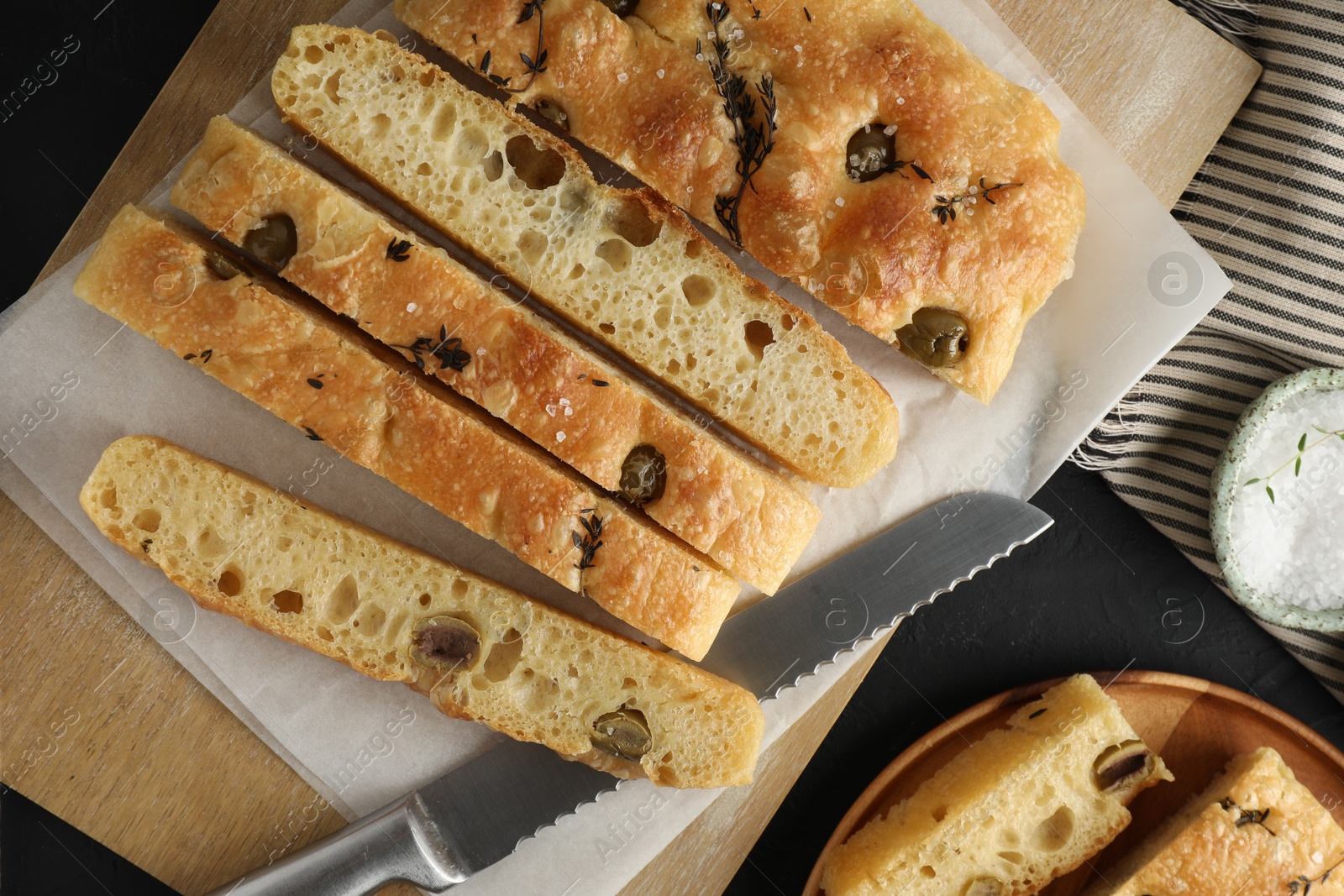 Photo of Slices of delicious focaccia bread with olives, thyme, salt and knife on black table, flat lay