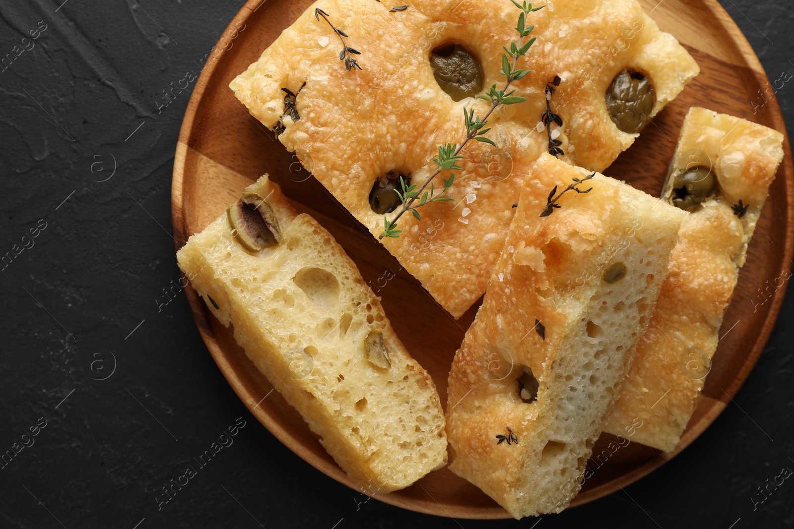 Photo of Slices of delicious focaccia bread with olives, thyme and salt on black table, top view