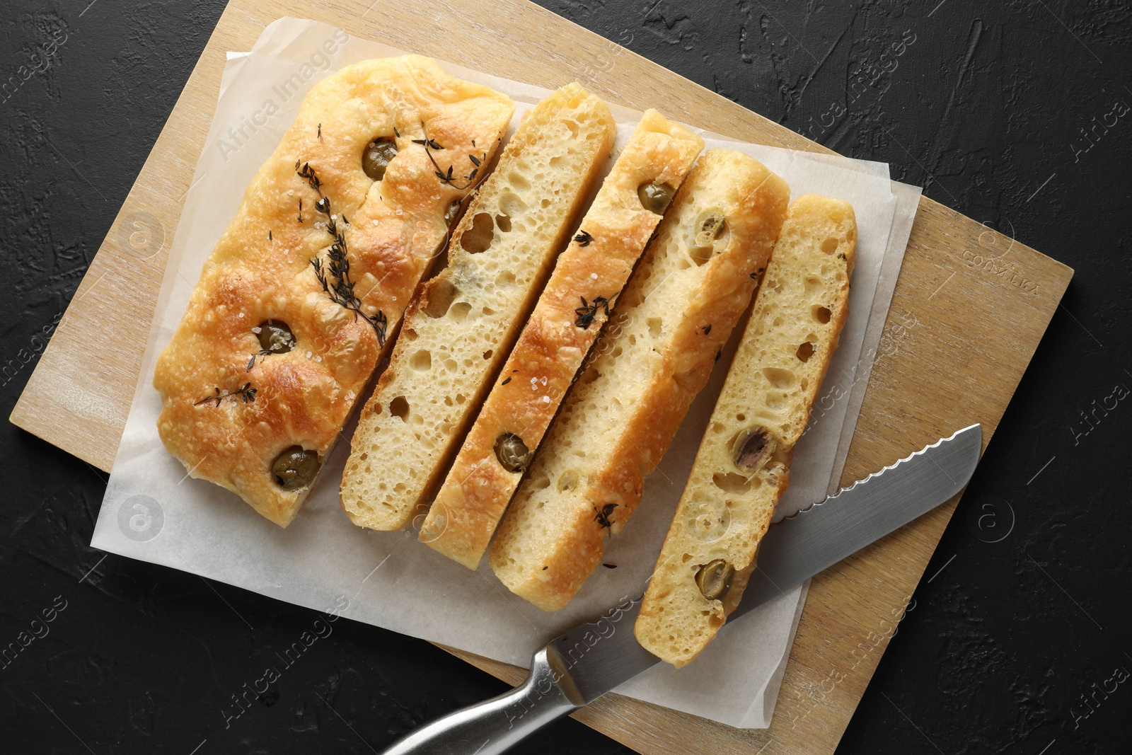 Photo of Slices of delicious focaccia bread with olives, thyme, salt and knife on black table, top view