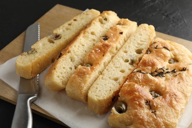 Photo of Slices of delicious focaccia bread with olives, thyme, salt and knife on black table, closeup