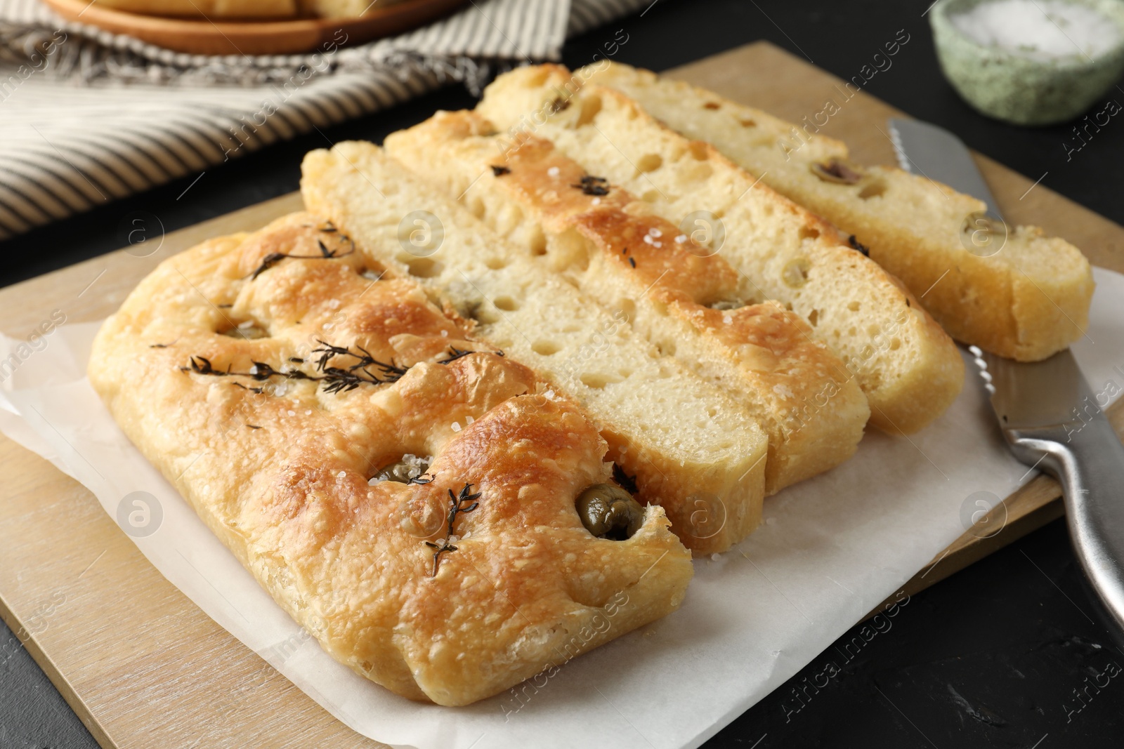 Photo of Slices of delicious focaccia bread with olives, thyme, salt and knife on black table, closeup