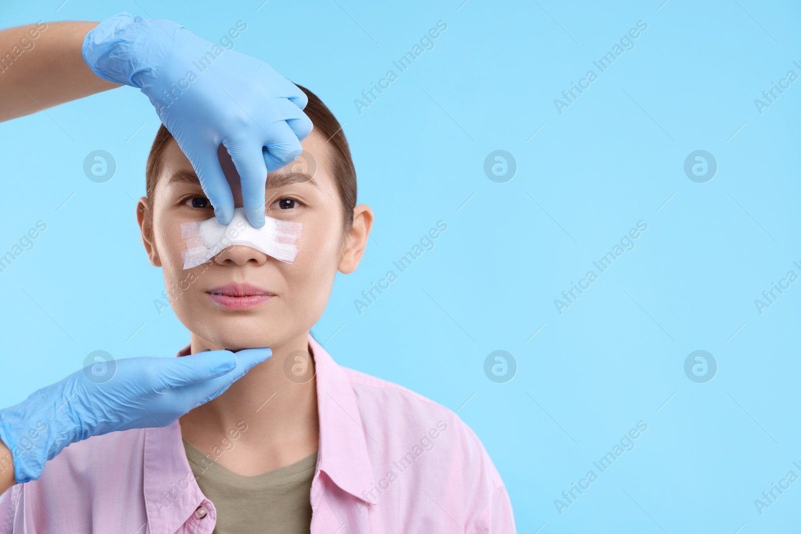 Photo of Doctor checking patient's nose after plastic surgery operation on light blue background, closeup. Space for text