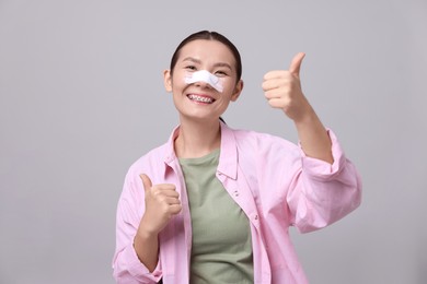 Photo of Woman with medical bandage on her nose after plastic surgery operation showing thumbs up against light grey background