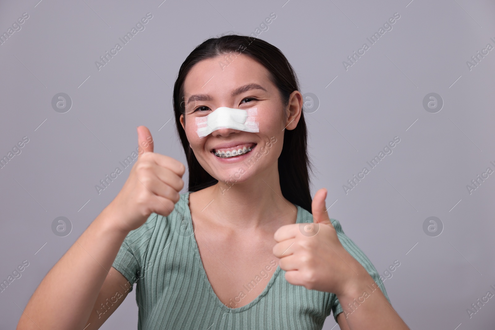 Photo of Woman with medical bandage on her nose after plastic surgery operation showing thumbs up against light grey background