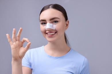 Photo of Woman with medical bandage on her nose after plastic surgery operation showing ok gesture against grey background