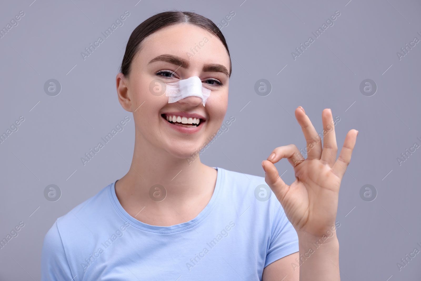 Photo of Woman with medical bandage on her nose after plastic surgery operation showing ok gesture against grey background