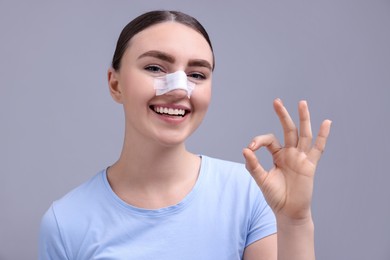 Photo of Woman with medical bandage on her nose after plastic surgery operation showing ok gesture against grey background