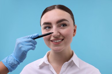 Photo of Doctor drawing marks on patient's nose for plastic surgery operation against light blue background, closeup