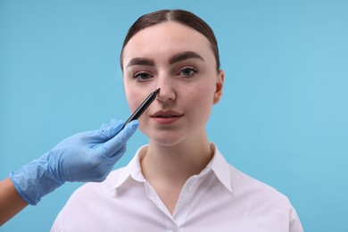 Photo of Doctor drawing marks on patient's nose for plastic surgery operation against light blue background, closeup