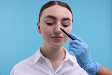 Photo of Doctor drawing marks on patient's nose for plastic surgery operation against light blue background, closeup