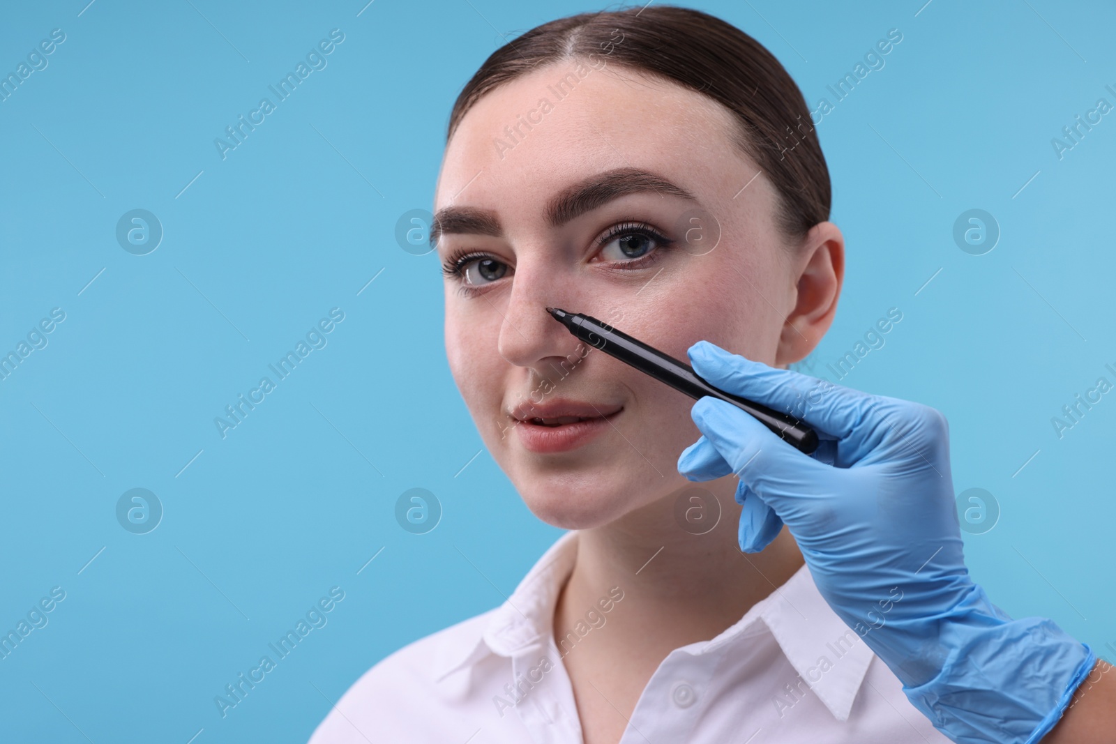 Photo of Doctor drawing marks on patient's nose for plastic surgery operation against light blue background, closeup