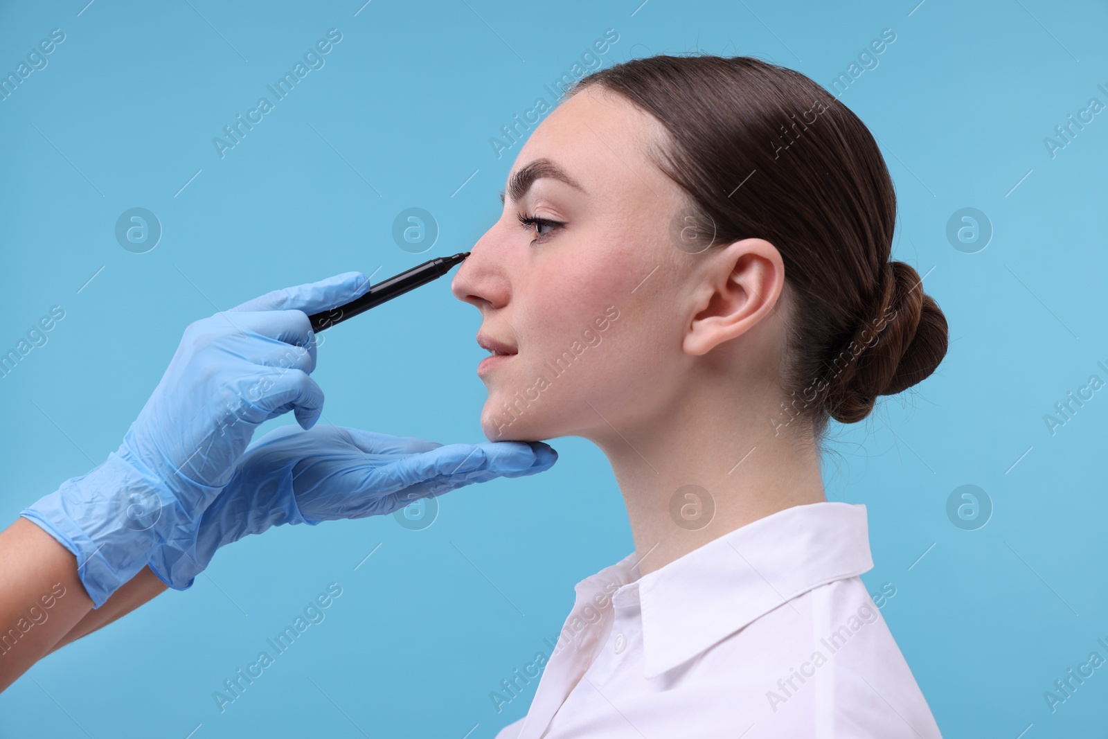 Photo of Doctor drawing marks on patient's nose for plastic surgery operation against light blue background, closeup